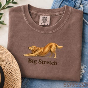May include: A brown crew neck t-shirt featuring a graphic of a golden-colored dog in a stretching pose. Below the dog, the text reads Big Stretch in a dark brown font. The shirt has a Comfort Colors brand label on the inside of the collar.