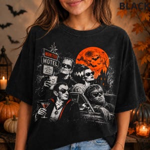May include: A black t-shirt featuring a graphic print of classic horror movie characters including Frankenstein, his bride, Dracula, and a werewolf. The design includes a "Monster Motel" sign, a large orange moon, and bats against a dark background.