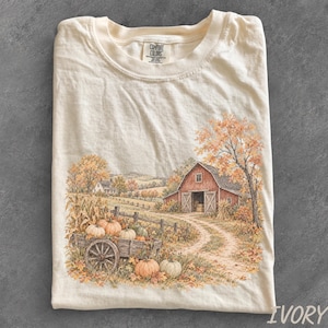 May include: An ivory short-sleeve t-shirt featuring a detailed graphic print of a rural autumn scene. The illustration depicts a red barn, a wooden wagon filled with orange pumpkins, corn stalks, and trees with fall foliage along a dirt path.