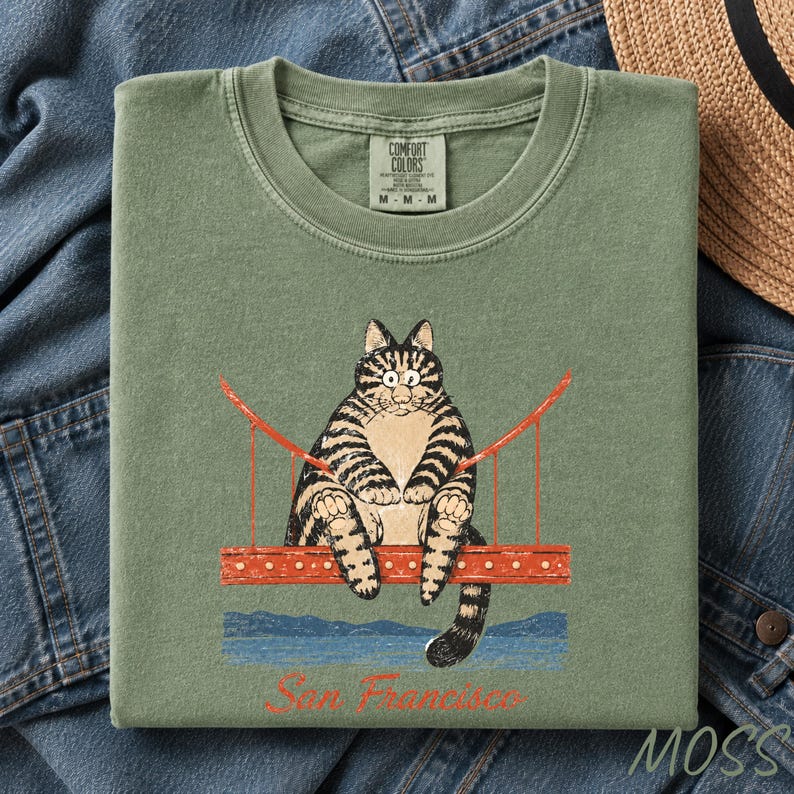 May include: A sage green crewneck t-shirt featuring a graphic of a tabby cat sitting on a red bridge. Below the bridge is a blue water illustration and the red text San Francisco. The inner neck label reads Comfort Colors size M.