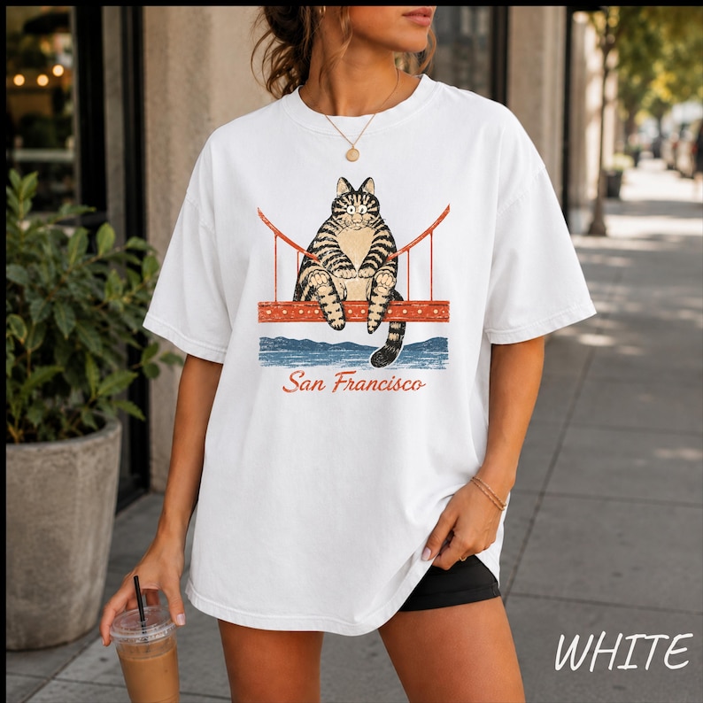 May include: A white short-sleeve t-shirt featuring a graphic of a brown tabby cat sitting on the Golden Gate Bridge. Below the bridge is a blue water illustration and the red text San Francisco. The shirt has a crew neckline and loose fit.