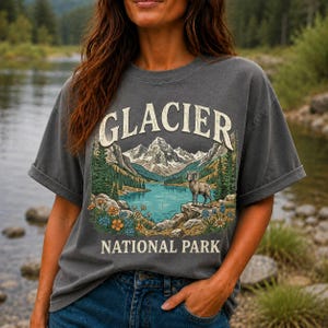 Może przedstawiać: Ciemnoszary t-shirt z krótkim rękawem z nadrukiem graficznym przedstawiającym Park Narodowy Glacier. Wzór zawiera pasmo górskie, jezioro, owcę kanadyjską i polne kwiaty oraz napis GLACIER NATIONAL PARK wykonany pogrubioną czcionką.