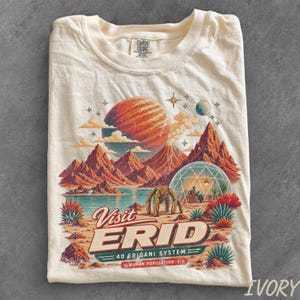 May include: An ivory t-shirt featuring a graphic print of a landscape with red mountains, a large planet, a geodesic dome, and a robotic figure. The text reads Visit ERID 40 Eridani System Human Population: 1. The shirt has a Comfort Colors brand label.
