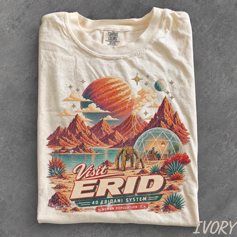 May include: An ivory t-shirt featuring a graphic print of a landscape with red mountains, a large planet, a geodesic dome, and a robotic figure. The text reads Visit ERID 40 Eridani System Human Population: 1. The shirt has a Comfort Colors brand label.