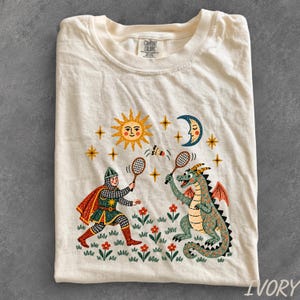 May include: An ivory t-shirt featuring a graphic print of a knight and a dragon playing badminton in a field of flowers. Above them are a yellow sun and a blue crescent moon with stars. The shirt has a Comfort Colors brand label at the neck.