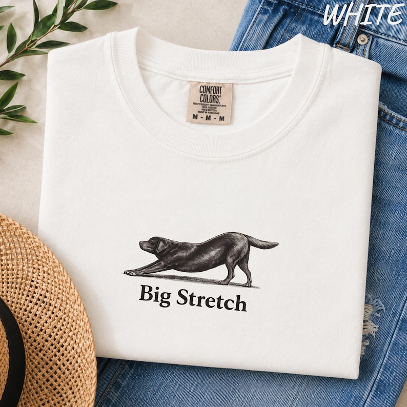 K&ouml;nnte beinhalten: Ein wei&szlig;es T-Shirt mit Rundhalsausschnitt und einer schwarz-wei&szlig;en Illustration eines schwarzen Labrador-Hundes in einer Dehnposition. Unter dem Hund ist der Text "Big Stretch" in einer schwarzen Serifenschrift gedruckt. Das Shirt hat ein Comfort Colors Etikett.