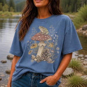 May include: A blue short-sleeve t-shirt featuring a graphic print of a toad sitting on a large mushroom, surrounded by wildflowers, a pinecone, and a small snail. The illustration is rendered in muted earth tones, contrasting with the blue fabric.