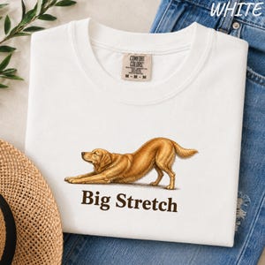 May include: A white t-shirt featuring a graphic of a golden-colored dog in a stretching pose. Below the dog, the text "Big Stretch" is printed in dark brown letters. The shirt has a Comfort Colors brand label on the inside of the collar.