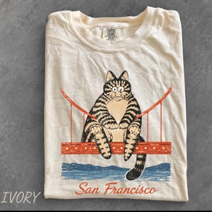 May include: An ivory t-shirt featuring a graphic of a tabby cat sitting on a red Golden Gate Bridge structure. Below the bridge is a blue water illustration and the text San Francisco. The shirt has a Comfort Colors brand label at the neck.
