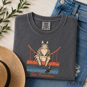 May include: A charcoal gray t-shirt featuring a graphic of a tabby cat sitting on a red Golden Gate Bridge structure. Below the bridge, the text San Francisco is printed in a red script font. The shirt has a crew neckline and a Comfort Colors label.