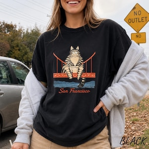May include: A black short-sleeve t-shirt featuring a graphic of a brown tabby cat sitting on a red bridge structure. Below the cat, the text San Francisco is printed in a cursive red font. The design includes blue water elements at the base.