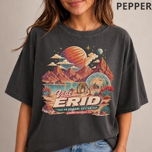May include: A charcoal gray t-shirt featuring a graphic print of a space landscape with red mountains, a large planet, and a domed structure. The text reads Visit ERID, 40 ERIDANI SYSTEM, HUMAN POPULATION: 1. The design includes a robotic figure.