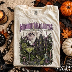 Könnte beinhalten: Ein cremefarbenes T-Shirt mit einem Grafikdruck und dem Text "Midnight Margaritas EST. 1998". Das Design zeigt ein Spukhaus, ein Margaritaglas mit Limettenscheibe, einen Vollmond, einen Frosch, eine Zaubertrankflasche und eine Kerze.