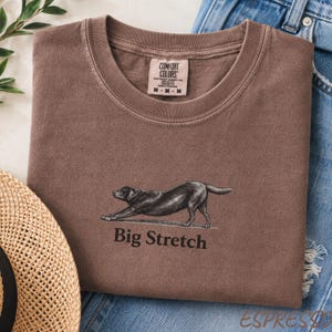 K&ouml;nnte beinhalten: Ein braunes T-Shirt mit Rundhalsausschnitt und einer Grafik eines schwarzen Hundes in Dehnposition &uuml;ber dem Text Big Stretch. Das T-Shirt hat ein Comfort Colors Markenetikett am Innenkragen und liegt neben blauem Jeansstoff und einem Strohhut.