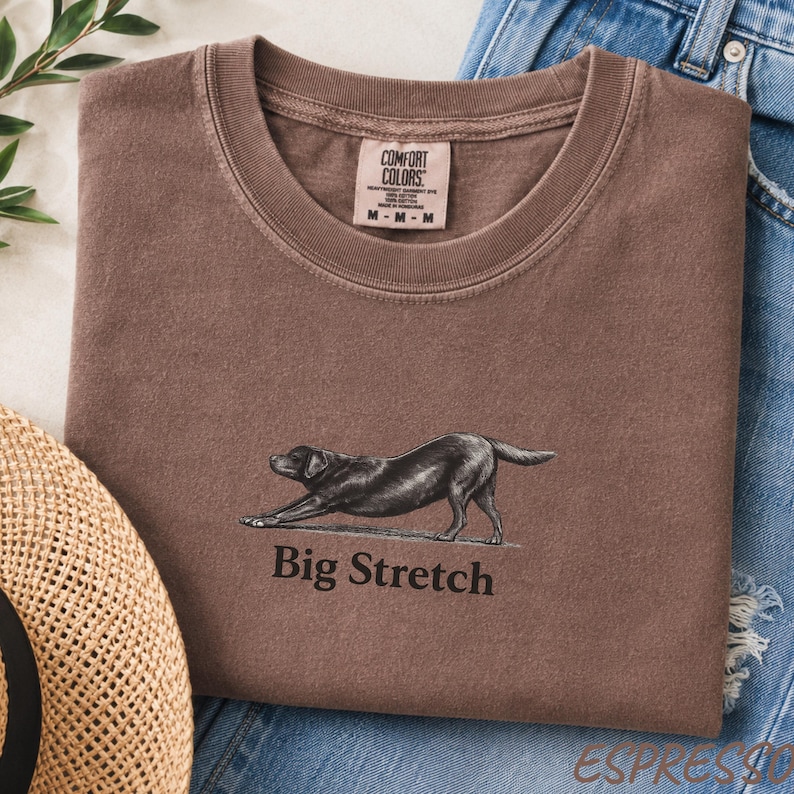 K&ouml;nnte beinhalten: Ein braunes T-Shirt mit Rundhalsausschnitt und einer Grafik eines schwarzen Hundes in Dehnposition &uuml;ber dem Text Big Stretch. Das T-Shirt hat ein Comfort Colors Markenetikett am Innenkragen und liegt neben blauem Jeansstoff und einem Strohhut.