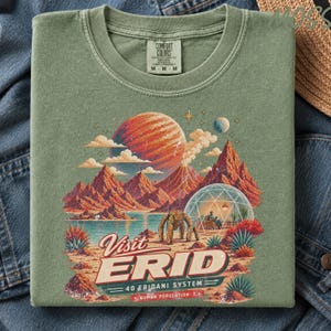 May include: A sage green t-shirt featuring a graphic print of a space landscape with red mountains, a large planet, and a domed habitat. The text reads Visit ERID 40 ERIDANI SYSTEM HUMAN POPULATION: 1. The shirt has a Comfort Colors brand label.