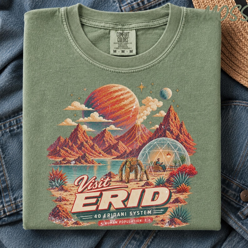 May include: A sage green t-shirt featuring a graphic print of a space landscape with red mountains, a large planet, and a domed habitat. The text reads Visit ERID 40 ERIDANI SYSTEM HUMAN POPULATION: 1. The shirt has a Comfort Colors brand label.