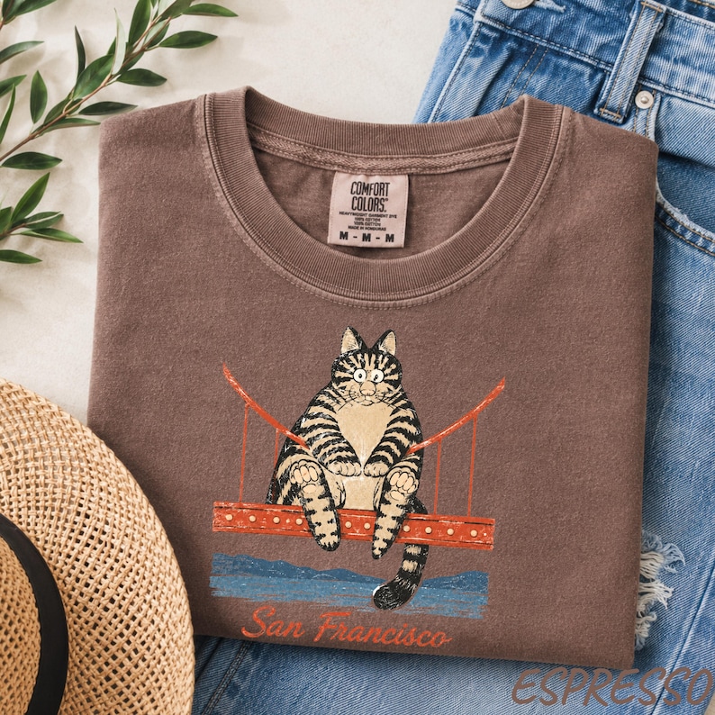 May include: A brown crew neck t-shirt featuring a graphic of a tabby cat sitting on a red bridge structure. Below the bridge, the text San Francisco is printed in a cursive font. The shirt has a Comfort Colors brand label on the inner neckline.