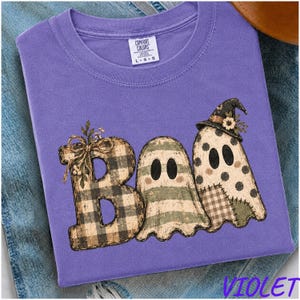 May include: A purple crew neck t-shirt featuring a graphic of the word "BOO" where the letter B is a plaid pattern with a bow, and the two O's are stylised ghosts with one wearing a witch hat. The shirt has a Comfort Colors brand label.
