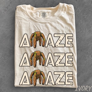 May include: An ivory-colored t-shirt featuring three stacked rows of the word AMAZE in a bold, outlined font. The letter M in each row is replaced by a brown, rounded, multi-legged robotic figure with small green accents on its body.