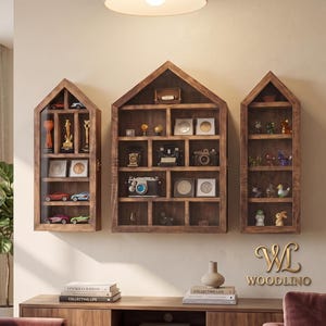 May include: Three house-shaped wooden display shelves with glass fronts mounted on a wall. The shelves feature multiple compartments containing various small collectibles, including toy cars, cameras, coins, and figurines. The logo WOODLINO is visible.