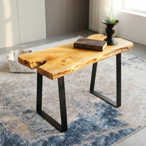 May include: A rectangular bench featuring a natural live-edge light wood top with visible grain and knots, supported by two black metal U-shaped legs. A dark brown book and a black vase with a small plant are placed on the wooden surface.