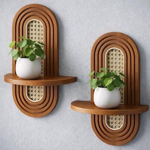 May include: Two wooden wall shelves with a natural finish, each featuring a curved design with a woven cane insert. Each shelf holds a white ceramic pot with a green leafy plant. The shelves are mounted on a light gray wall.