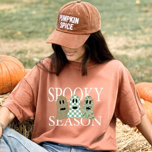 Op de afbeelding: Een roestkleurig t-shirt met de witte tekst SPOOKY SEASON en drie spookillustraties met luipaard-, ruit- en camouflagepatronen. Een bijpassende roestkleurige corduroy baseballpet met de witte tekst PUMPKIN SPICE.