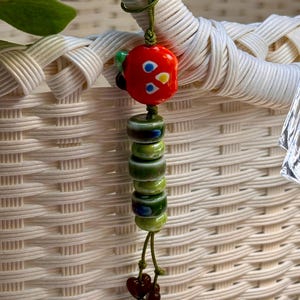 May include: A decorative charm featuring a bright red glass bead with blue and yellow accents at the top, followed by a vertical row of five cylindrical green glass beads with dark markings, ending with two small dark brown heart-shaped beads.
