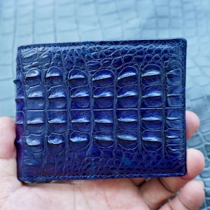 Puede incluir: Una cartera plegable de cuero azul profundo con una textura de piel de cocodrilo en relieve. La superficie muestra un patrón de escamas distintivo y repetitivo con un acabado brillante, exhibiendo un rico degradado de tonos azules oscuros.