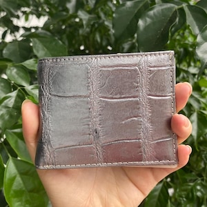 May include: A metallic silver bifold wallet featuring a textured crocodile skin pattern. The wallet has a rectangular shape with visible stitching along the edges and a glossy finish that reflects light across its surface.