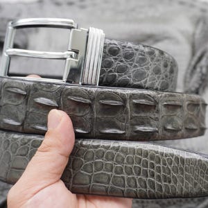 Op de afbeelding: Een grijze riem met een gestructureerd, verhoogd schubbenpatroon over de gehele band. De riem is voorzien van een gepolijste zilverkleurige metalen gesp met een rechthoekig frame en een geribbeld detail aan de zijkant, met een natuurlijke reptielhuid.