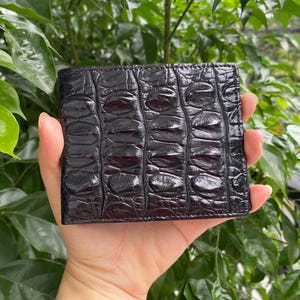 May include: A dark, glossy black bifold wallet featuring a prominent, raised, and textured crocodile skin pattern. The surface of the wallet displays a repeating, organic scale design with a polished finish that reflects light across its rectangular form.