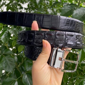 May include: A black belt with a textured, embossed pattern resembling reptile skin. The belt features a polished, silver-toned metal buckle with a rectangular frame and a single prong, providing a sleek and reflective contrast to the dark strap.
