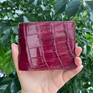 May include: A bifold wallet crafted from deep burgundy leather with a glossy, textured finish that mimics the appearance of crocodile skin. The wallet features visible stitching along the edges and a rectangular shape with a distinct scale pattern.