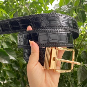 May include: A black belt with a glossy, textured crocodile-skin pattern and a polished gold-tone metal buckle. The belt features a stitched border along the edges and a rectangular frame buckle with a central prong, showcasing a shiny finish.