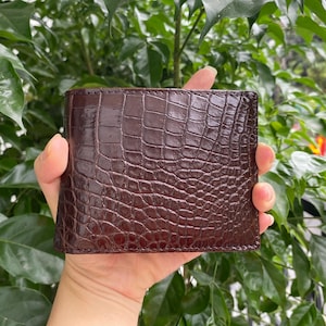 Op de afbeelding: Een donkerbruine bifold portemonnee met een glanzende, in reliëf gemaakte krokodillenleertextuur. Het rechthoekige accessoire heeft een gepolijste afwerking en een duidelijk schubbenpatroon over het gehele oppervlak in een diepe mahoniekleur.