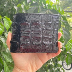 Puede incluir: Una cartera negra de dos pliegues con un patrón de escamas texturizado y elevado en la superficie exterior. La cartera tiene un acabado brillante y una forma rectangular con costuras visibles en los bordes, mostrando una textura orgánica distintiva.
