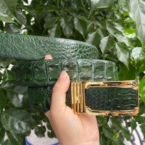 May include: A dark green belt featuring a textured, embossed pattern resembling reptile skin. The belt includes a rectangular gold-toned metal buckle with a polished finish and a central vertical bar design, showcasing a glossy, reflective surface.