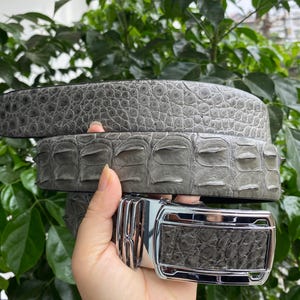 May include: A grey belt featuring a textured, scale-like pattern across the strap. The belt includes a polished silver-tone metal buckle with a rectangular frame and a central mechanism, showcasing a detailed, raised texture on the leather surface.