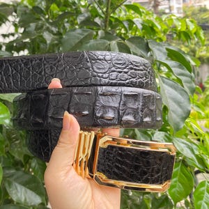 Op de afbeelding: Een zwarte riem met een getextureerd, reliëfpatroon dat lijkt op reptielenhuid. De riem heeft een rechthoekige, goudkleurige metalen gesp met een gepolijste afwerking en een centraal geometrisch ontwerp. De riem is opgerold en toont een consistent patroon.