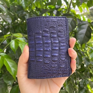 Può includere: Un portafoglio blu scuro caratterizzato da una texture in rilievo che imita la pelle di coccodrillo. Il portafoglio ha una forma rettangolare con cuciture visibili lungo i bordi e una finitura lucida che cattura la luce, mettendo in risalto le squame.