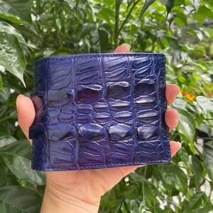 May include: A bi-fold wallet featuring a deep blue, glossy finish with a textured, raised scale pattern reminiscent of exotic skin. The wallet has visible stitching along the edges and a rectangular shape with a smooth, reflective surface texture.