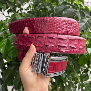 Puede incluir: Un cinturón de color rojo intenso con un patrón texturizado en relieve que imita la apariencia de escamas de reptil. El cinturón incluye una hebilla rectangular de metal plateado pulido con una incrustación roja a juego en el frente.