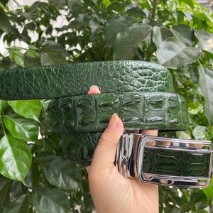 Puede incluir: Un cinturón de color verde oscuro con un patrón texturizado en relieve que imita la apariencia de la piel de reptil. El cinturón incluye una hebilla rectangular de metal plateado pulido con una incrustación verde a juego con la textura.