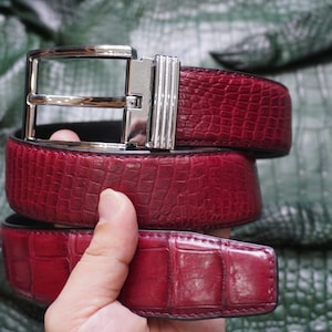 Op de afbeelding: Een dieprode riem met een textuur van krokodillenleer en een gepolijste zilverkleurige metalen gesp. De riem heeft een rechthoekige gesp met een verticaal geribbeld detail aan de zijkant, een glanzende afwerking en duidelijk zichtbare schubben.
