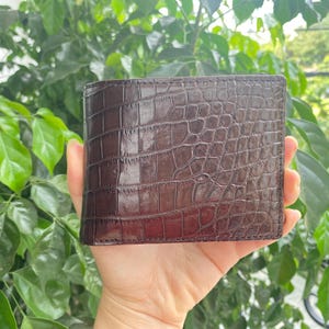 Op de afbeelding: Een donkerbruine bifold portemonnee met een glanzende, in reliëf aangebrachte krokodillenleertextuur. De portemonnee heeft een rechthoekige vorm met zichtbare stiksels langs de randen en toont een gedetailleerd schubbenpatroon over het gehele oppervlak.