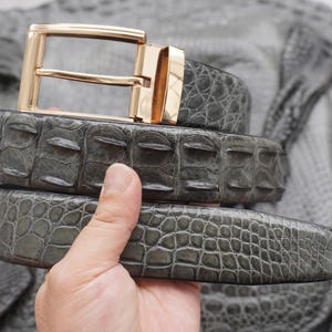 May include: A grey belt with a textured, scale-like pattern and a polished gold-tone metal buckle. The belt features a distinct, raised ridge design along the strap, showcasing a detailed, reptilian-inspired finish and a classic rectangular frame.