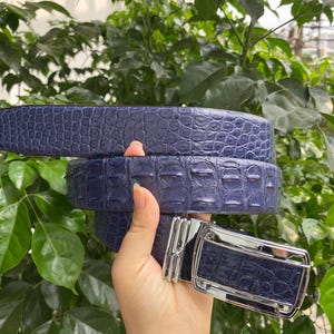 Puede incluir: Un cinturón azul oscuro con un patrón texturizado en relieve que imita la apariencia de piel exótica. El cinturón incluye una hebilla de metal plateado pulido con un marco rectangular y un mecanismo deslizante central para un cierre seguro.