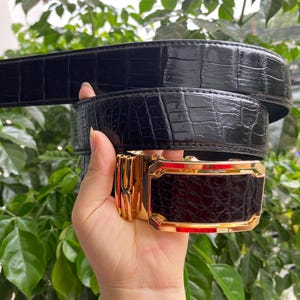 Könnte beinhalten: Ein schwarzer Gürtel mit glänzender Krokodilleder-Textur und einer rechteckigen goldfarbenen Metallschnalle. Die Schnalle hat ein poliertes Finish mit einem zentralen Einsatz, der dem schwarzen Krokodil-Prägemuster des Lederriemens entspricht.